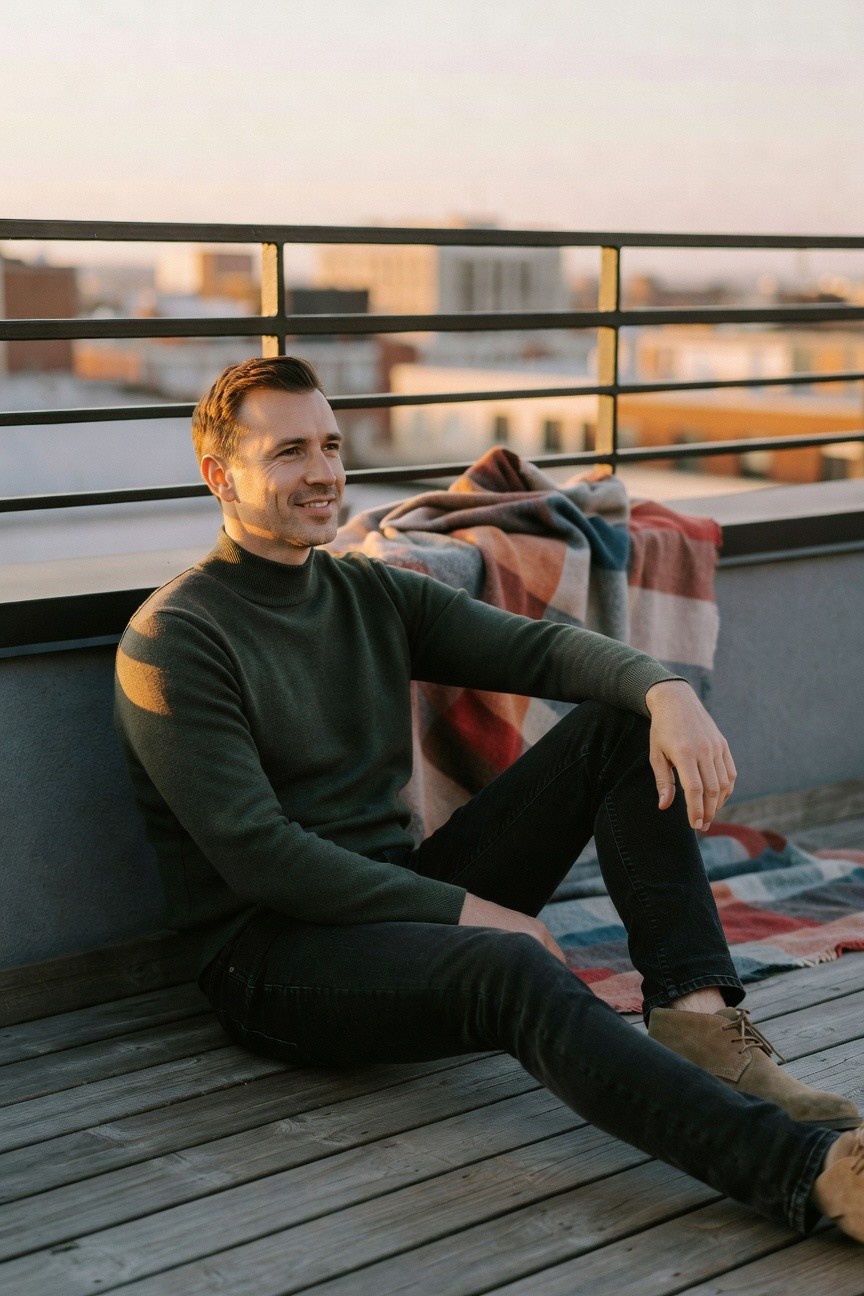 Man sitting casually in olive green turtleneck sweater, black slim jeans, and tan suede boots on wooden deck with plaid blanket nearby