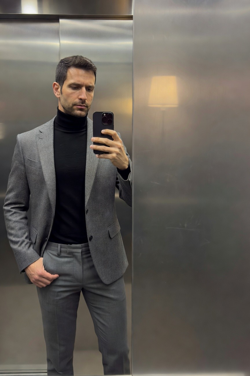 Man taking a mirror selfie in an elevator, wearing a textured grey blazer over a black turtleneck, slim grey pants, holding an iPhone, with a watch on his wrist