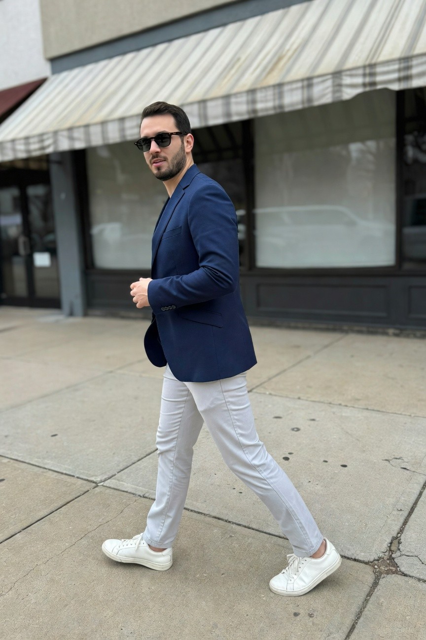 Man in fitted navy blue blazer, light grey slim pants, and white sneakers walking casually, side profile with sunglasses and trimmed beard