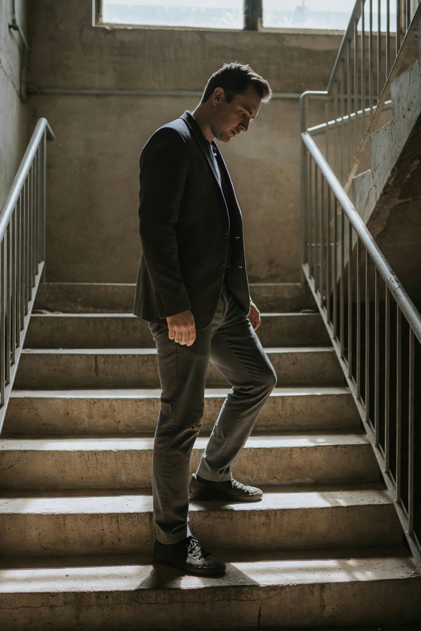 Man in dark blazer, slim grey pants, and black high-top sneakers ascending concrete stairs in an industrial stairwell, side profile view