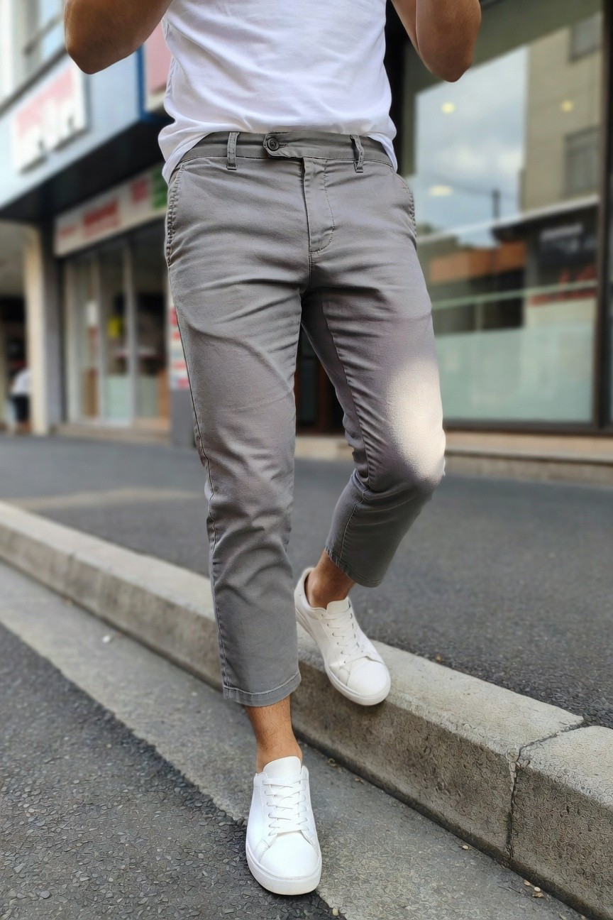 Man in slim grey ankle-length chinos, white t-shirt tucked at waist, and white sneakers, standing casually on a street curb