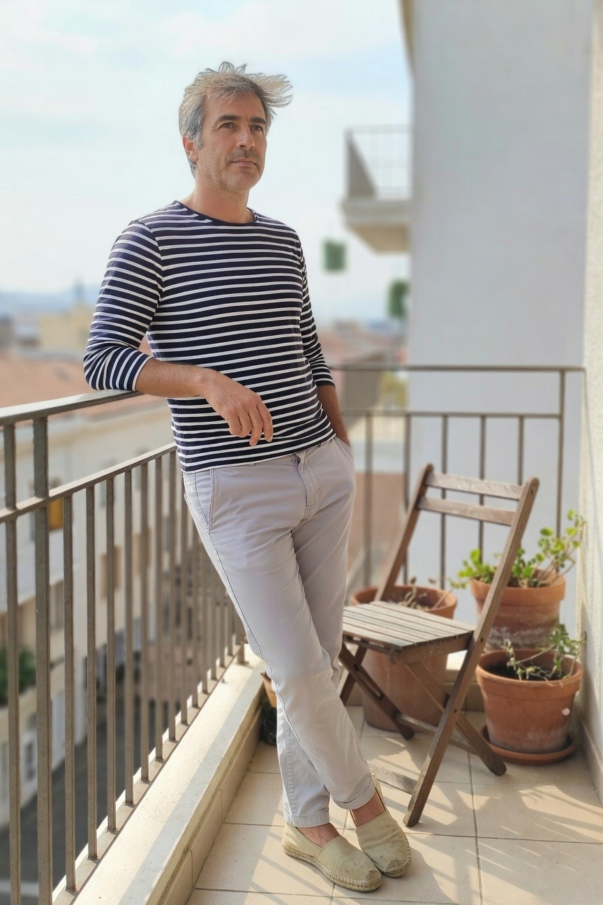 Man in navy and white horizontal striped long-sleeve cotton shirt, light grey slim-fit trousers, tan espadrilles, hands in pockets leaning against balcony rail with casual posture