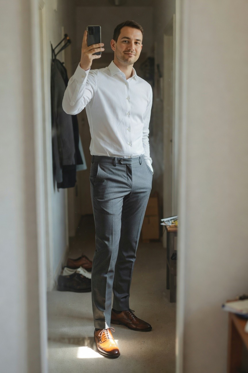 A man stands in front of a mirror wearing a white long-sleeve button-up shirt tucked into slim grey trousers, brown wingtip brogue shoes on his feet, holding a phone for a selfie with one hand in his trouser pocket.