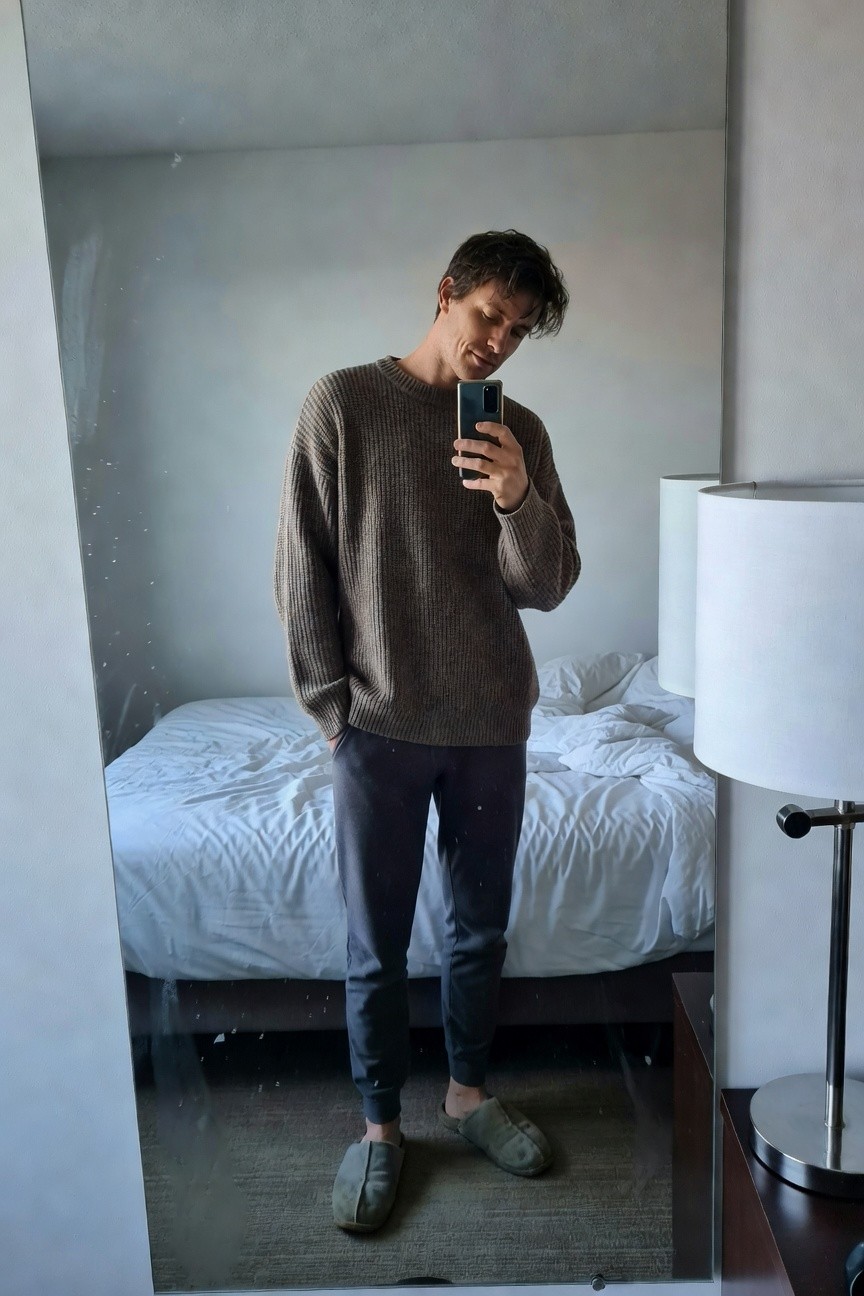 Mirror selfie of a man in an oversized camel-colored ribbed knit sweater, dark grey tapered joggers, and fuzzy slippers, standing in a simple bedroom with white bed and lamp visible