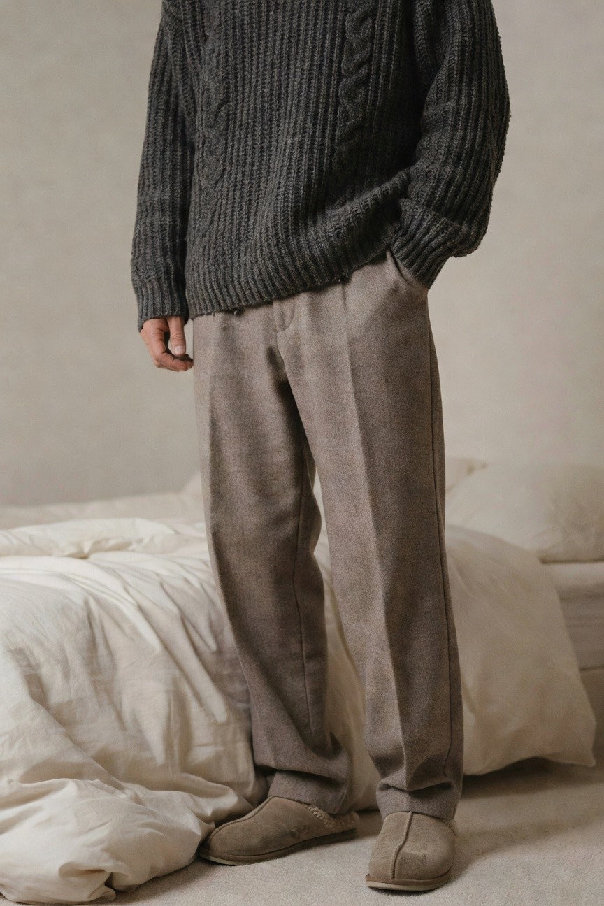 Man in chunky gray cable knit sweater, baggy taupe wool trousers, and beige shearling slippers standing by a white bed, relaxed indoor outfit