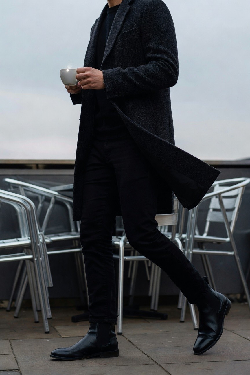 Man in slim black jeans, black knit top, long dark gray wool overcoat, and black Chelsea boots, holding a white mug while walking on a terrace with metal chairs nearby