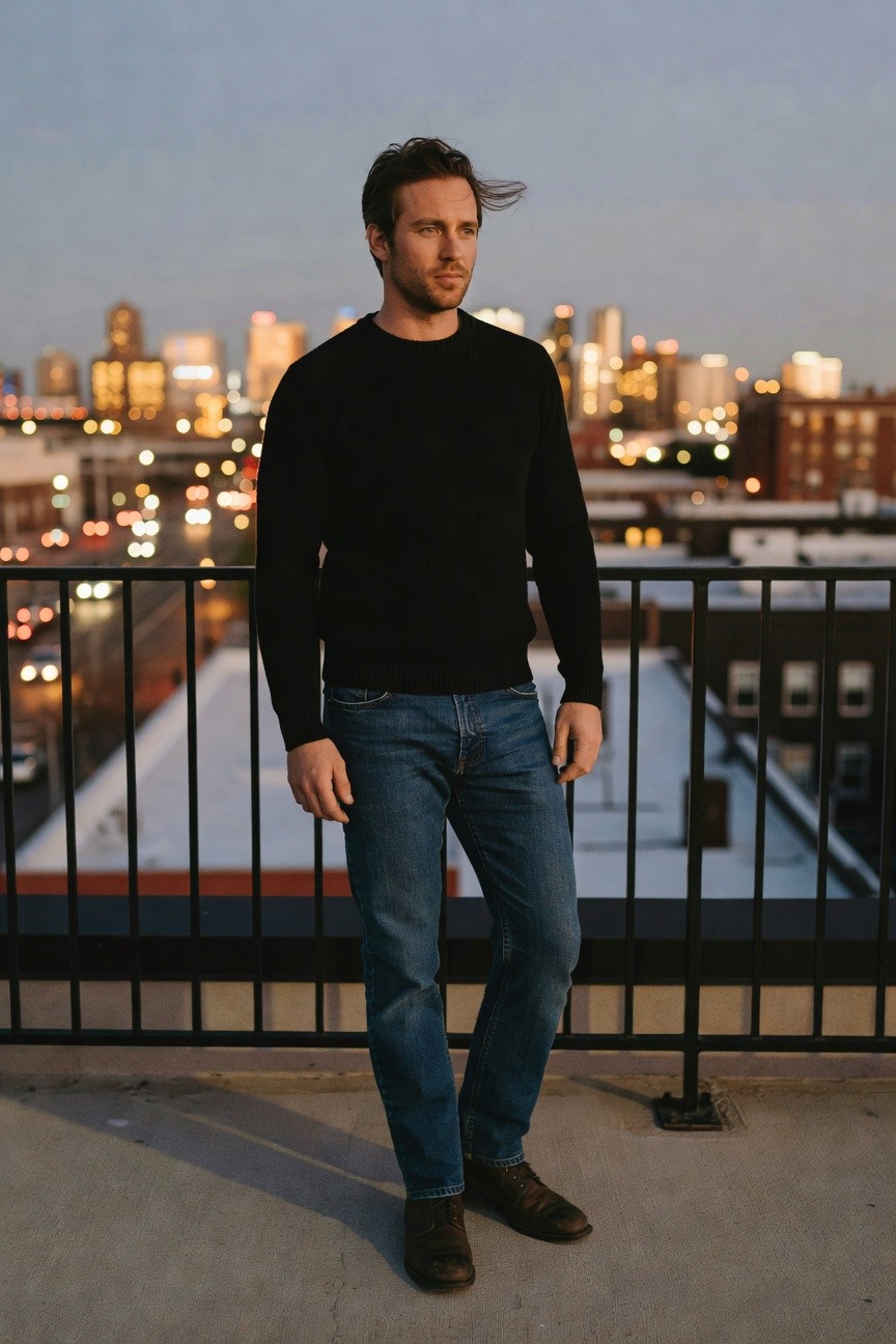 Man in black crewneck sweater, medium blue straight-leg jeans, and brown leather boots standing casually on a rooftop railing with city skyline in background at dusk