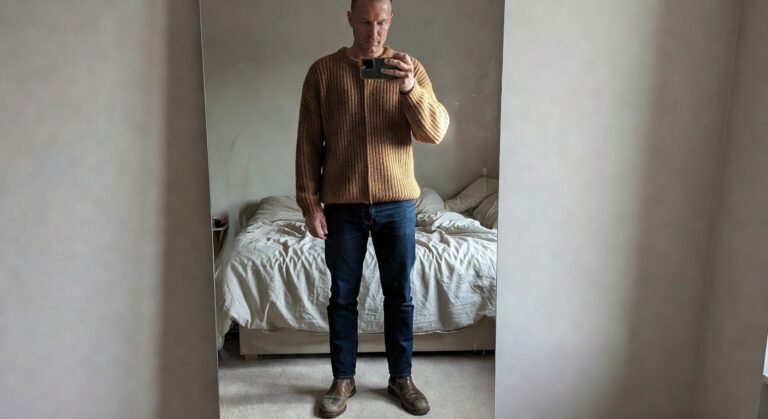 Man taking mirror selfie in open tan knit shawl-collar cardigan over white tee with dark straight-leg jeans and brown leather chelsea boots standing in bedroom
