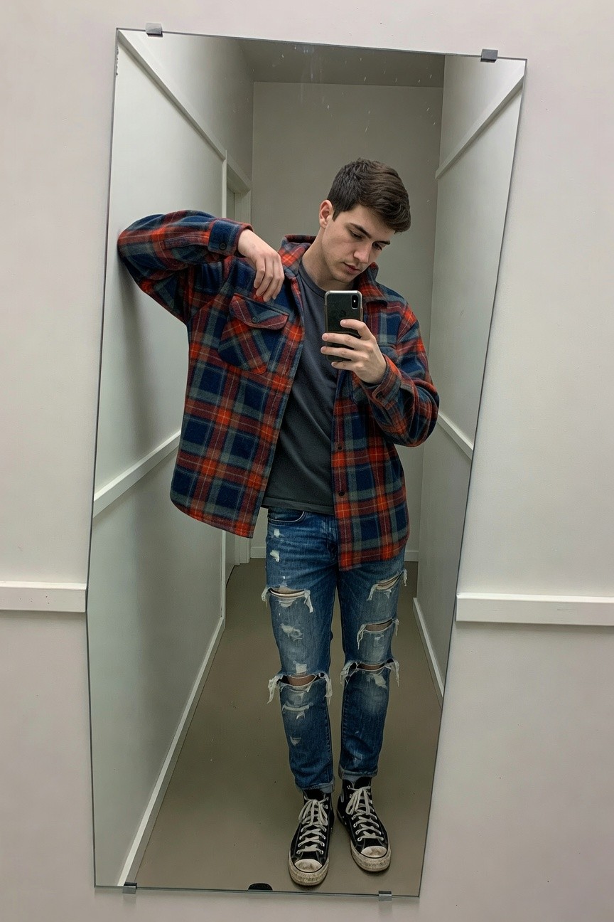 Young man posing in a full-length mirror wearing an open red-and-blue plaid flannel shirt over a gray t-shirt, distressed blue jeans with rips at knees and thighs, and black high-top Converse sneakers