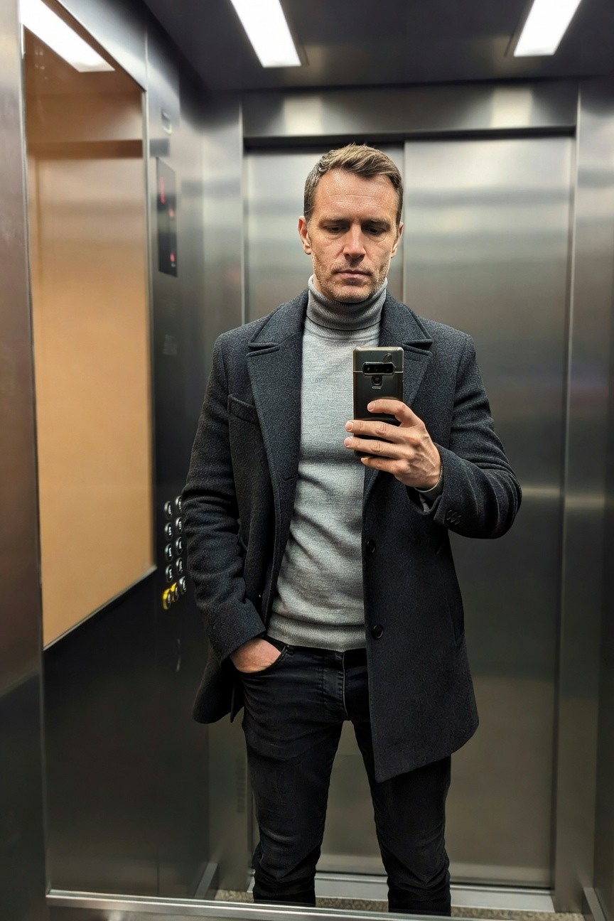 Man in elevator mirror selfie wearing slim dark jeans, light grey turtleneck sweater, charcoal wool overcoat with hands in pockets, holding phone, modern layered fall outfit