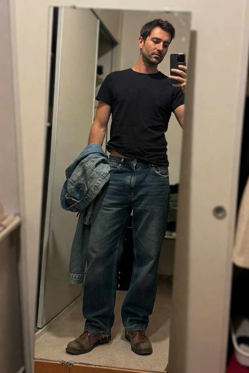 Mirror selfie of a man in a black short-sleeve t-shirt, baggy light blue jeans, a denim jacket draped over one shoulder, and brown leather work boots standing in a bedroom closet area