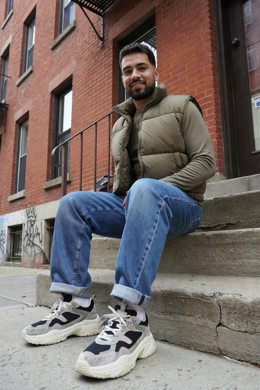 A man with a beard sits on urban steps wearing light blue baggy jeans cuffed at the ankles, an olive green quilted puffer vest over a light long-sleeve shirt, grey-and-white chunky sneakers, and a small black crossbody bag, against a brick building backdrop