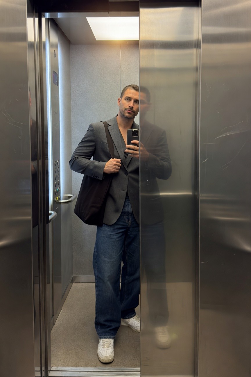 Man taking elevator selfie in open gray blazer over tee, black shoulder tote bag, dark baggy jeans, and white sneakers, with mirrored reflection visible