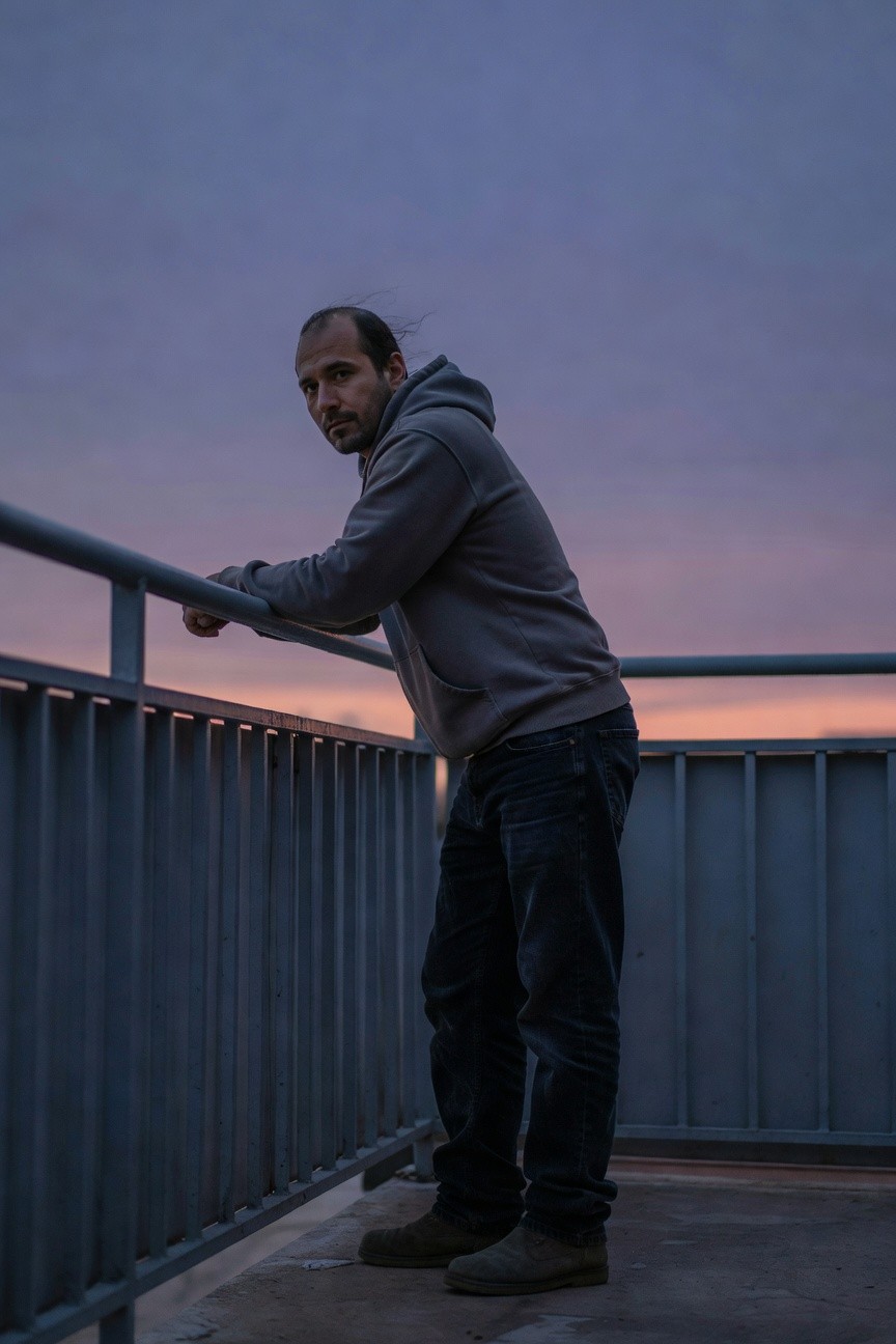 Man in grey hoodie, dark baggy jeans, and brown boots leaning casually on a metal railing at dusk