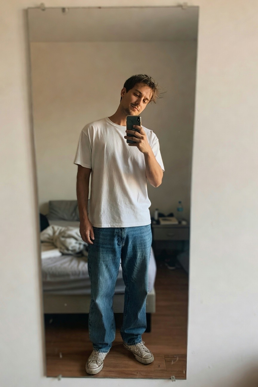 Man taking a mirror selfie in a bedroom, wearing an oversized white short-sleeve t-shirt, baggy mid-blue jeans, and white low-top sneakers, with tousled hair and subtle stubble