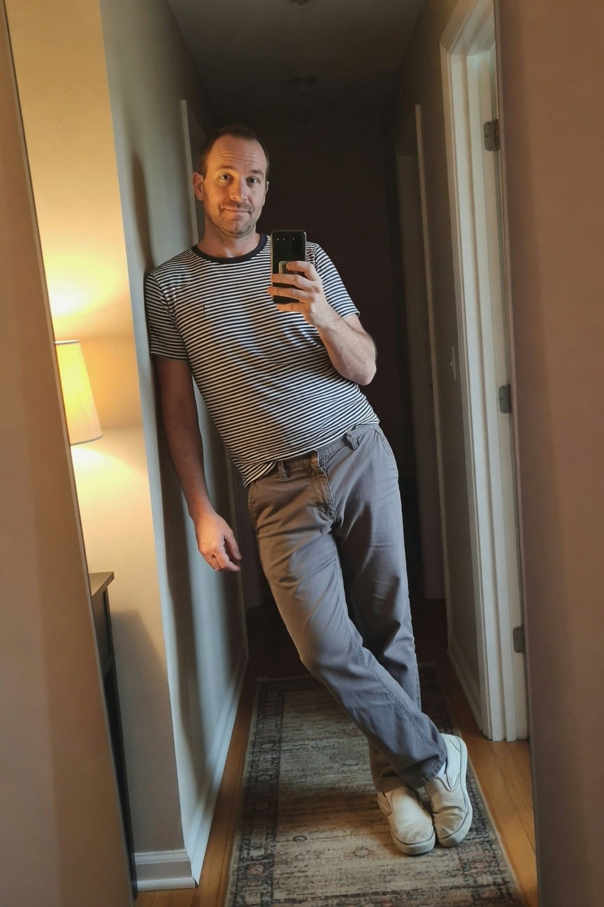 Man taking mirror selfie in narrow hallway, wearing black-and-white striped short-sleeve shirt, relaxed grey chinos, and white slip-on sneakers, leaning casually against wall