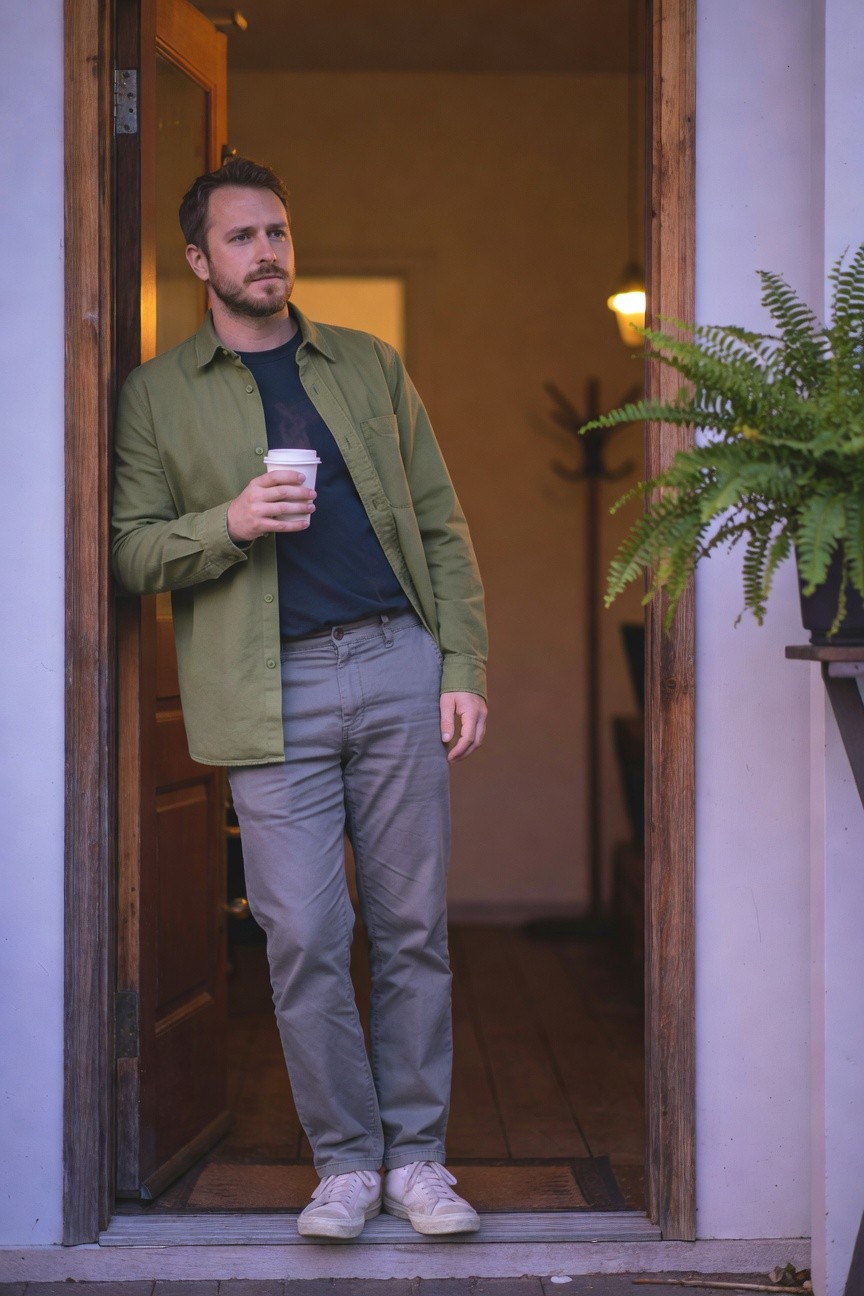 Man leaning in open doorway wearing open olive green button-up shirt over black t-shirt, relaxed grey chinos, and white sneakers, holding a white coffee cup