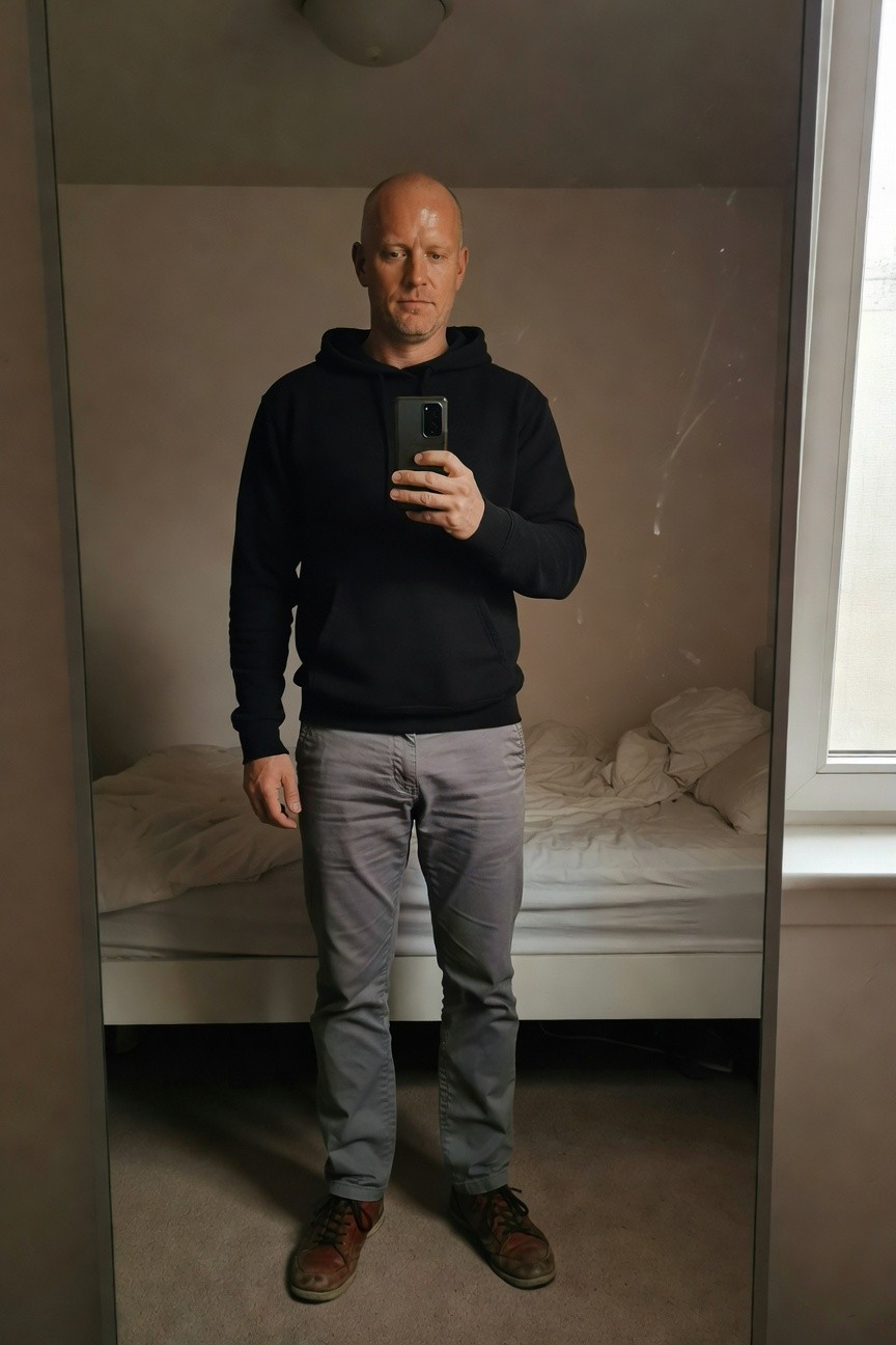 Bald man in black hoodie, grey chinos, and brown boots standing in bedroom mirror for casual selfie