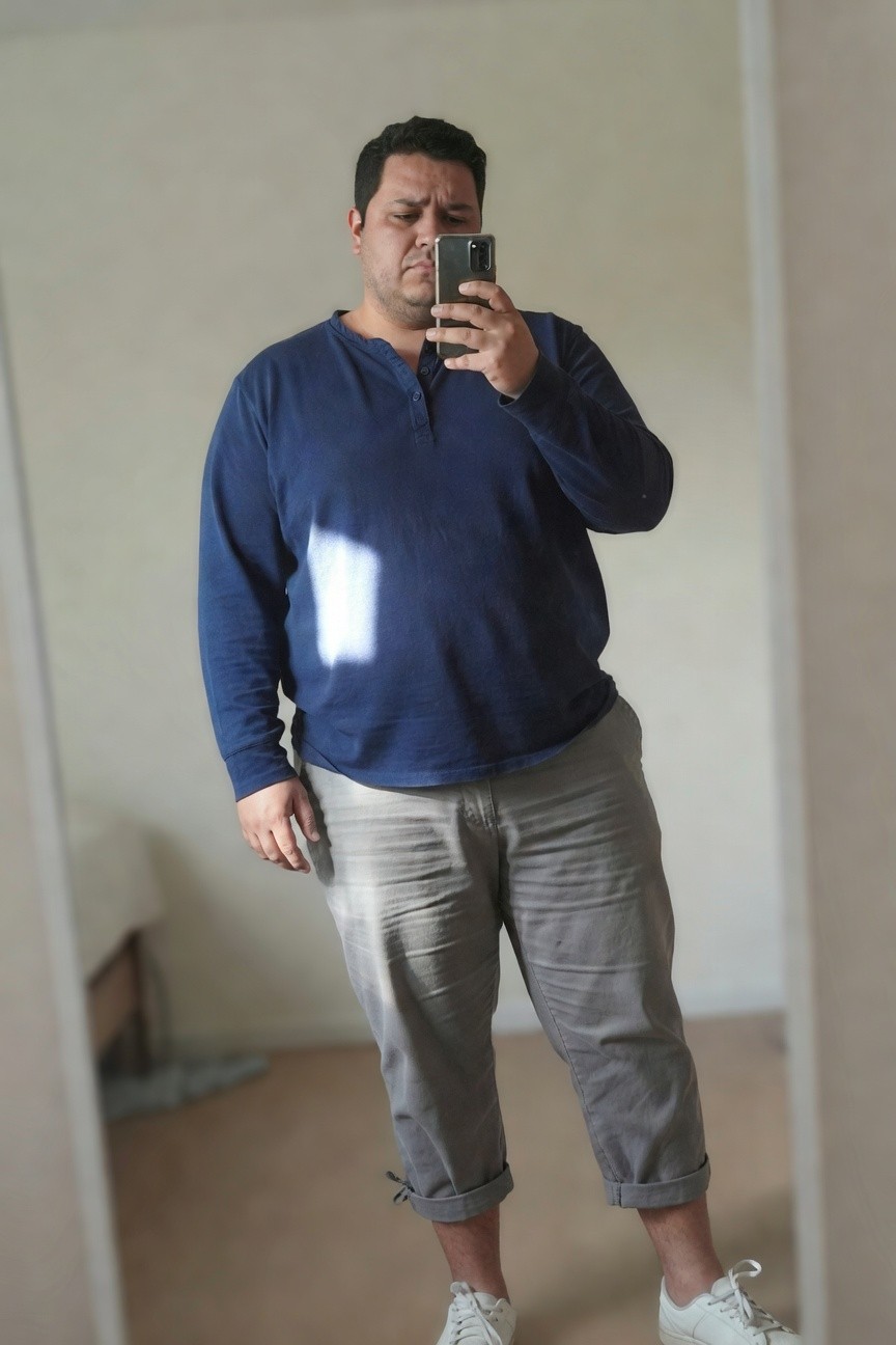Plus-size man in a navy long-sleeve henley shirt, cropped grey chinos, and white sneakers, taking a mirror selfie in a bedroom.