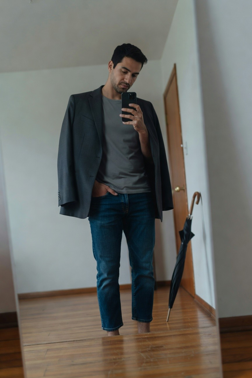 Mirror selfie of a man in an open dark gray blazer over a gray t-shirt, slim blue jeans cropped at the ankles showing bare feet, holding a phone, black umbrella propped nearby on wooden floor