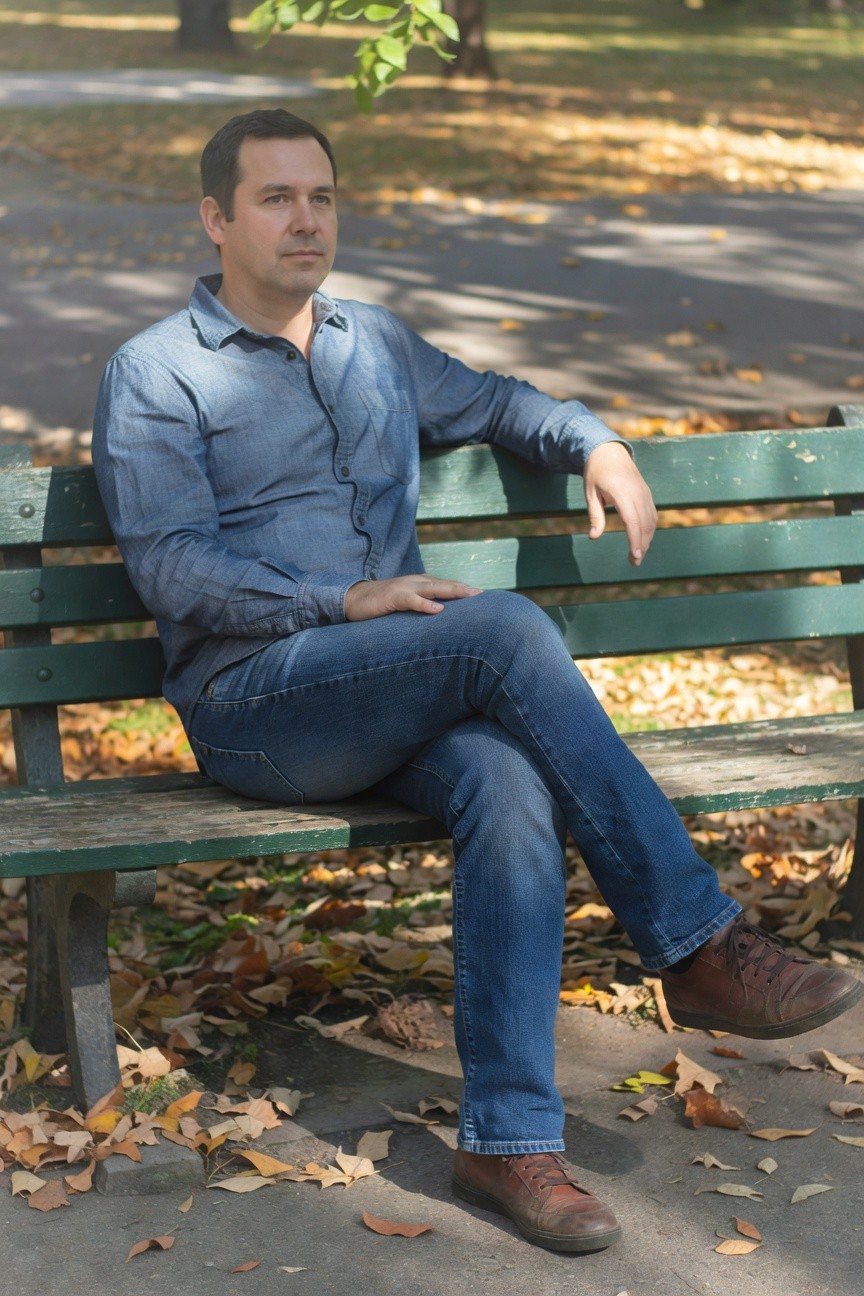 Man seated on park bench in light blue chambray long-sleeve shirt with sleeves rolled up, blue jeans, and reddish-brown leather boots, autumn leaves scattered nearby