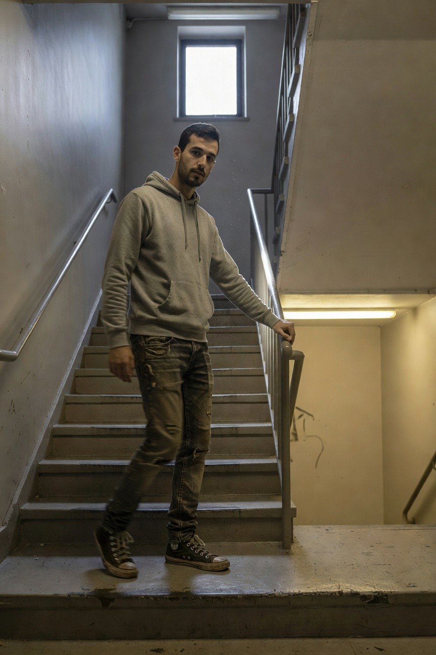 Man in light gray hoodie, dark distressed jeans, and black Converse high-tops standing on concrete stairs with hand on metal railing