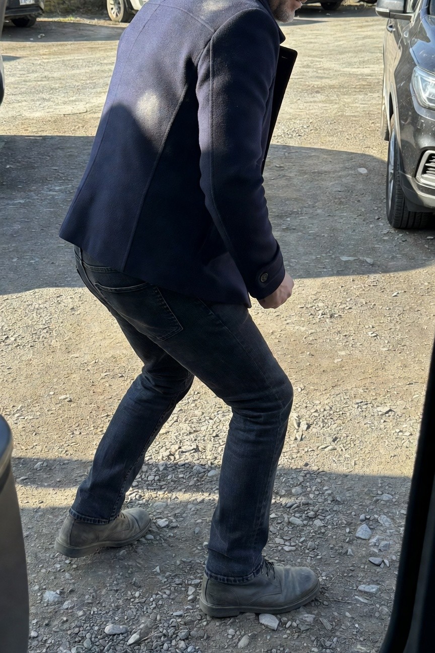 Side view of man in navy textured wool blazer, slim dark jeans, and gray suede lace-up boots standing near cars on gravel lot