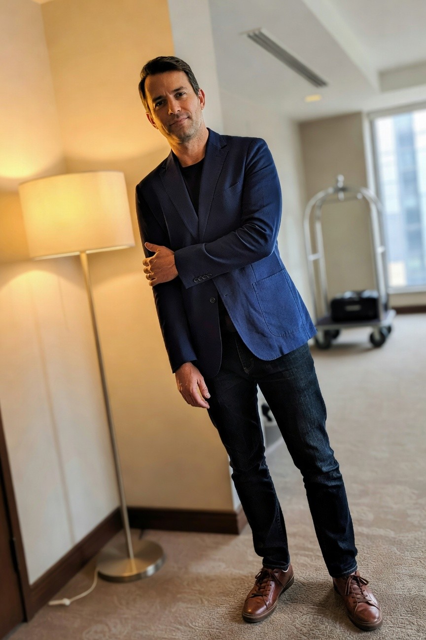 Man in navy textured blazer over black t-shirt, slim dark jeans, and brown laced leather shoes, standing casually in modern hotel room with lamp and cart in background