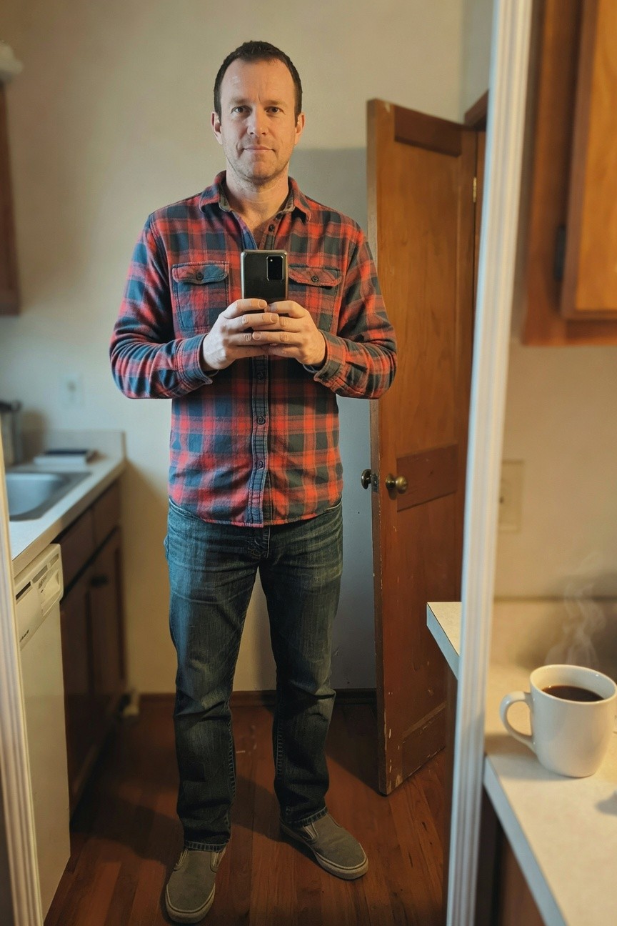 Man in red-black plaid flannel shirt, dark straight-leg jeans, and gray slip-on shoes, standing casually in a kitchen for a mirror selfie holding his phone