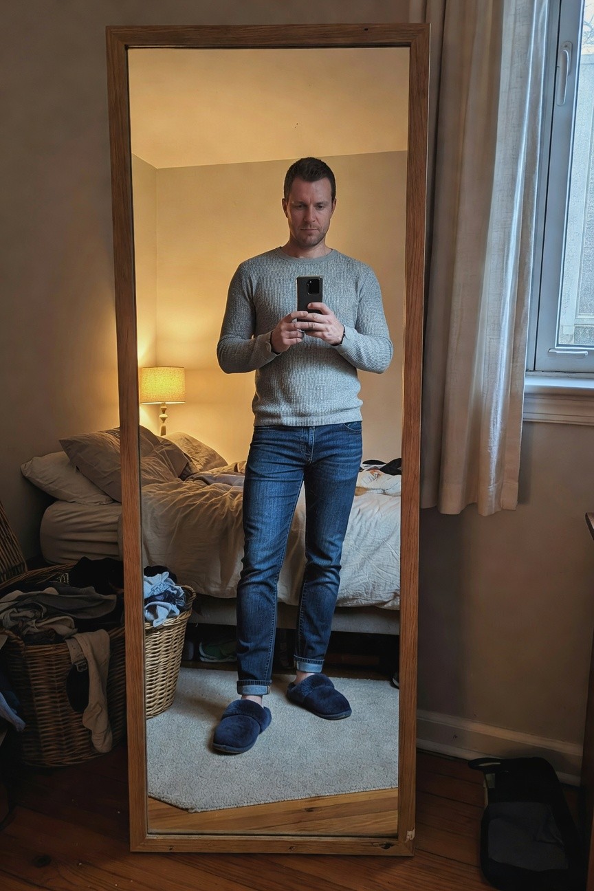 Man standing in front of mirror wearing light gray textured knit sweater, slim dark blue jeans cuffed at ankles, and fluffy blue slippers, holding phone for selfie in cozy bedroom setting