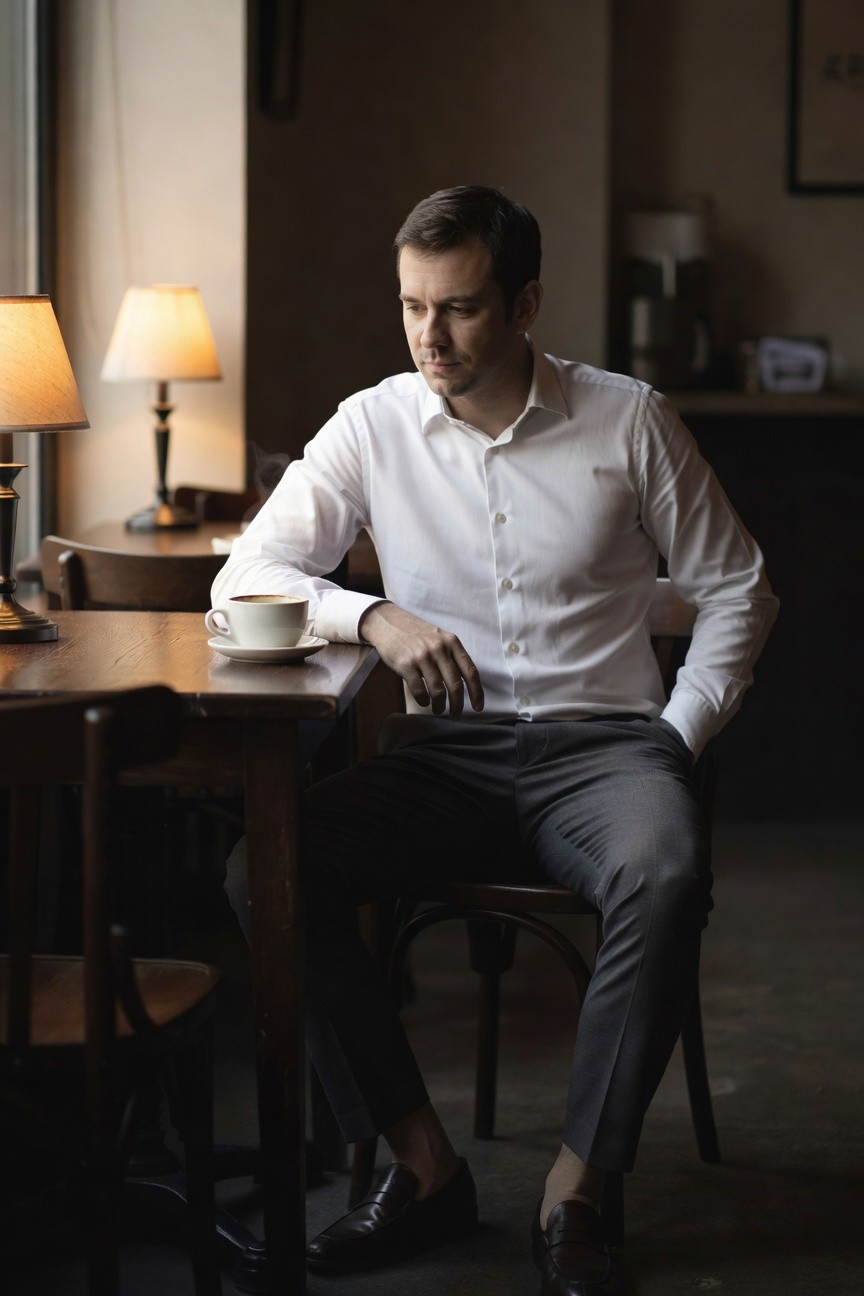 Man in minimalist outfit sitting at wooden cafe table, wearing long-sleeve white button-down shirt, slim grey dress pants, and brown loafers, holding coffee cup with thoughtful expression