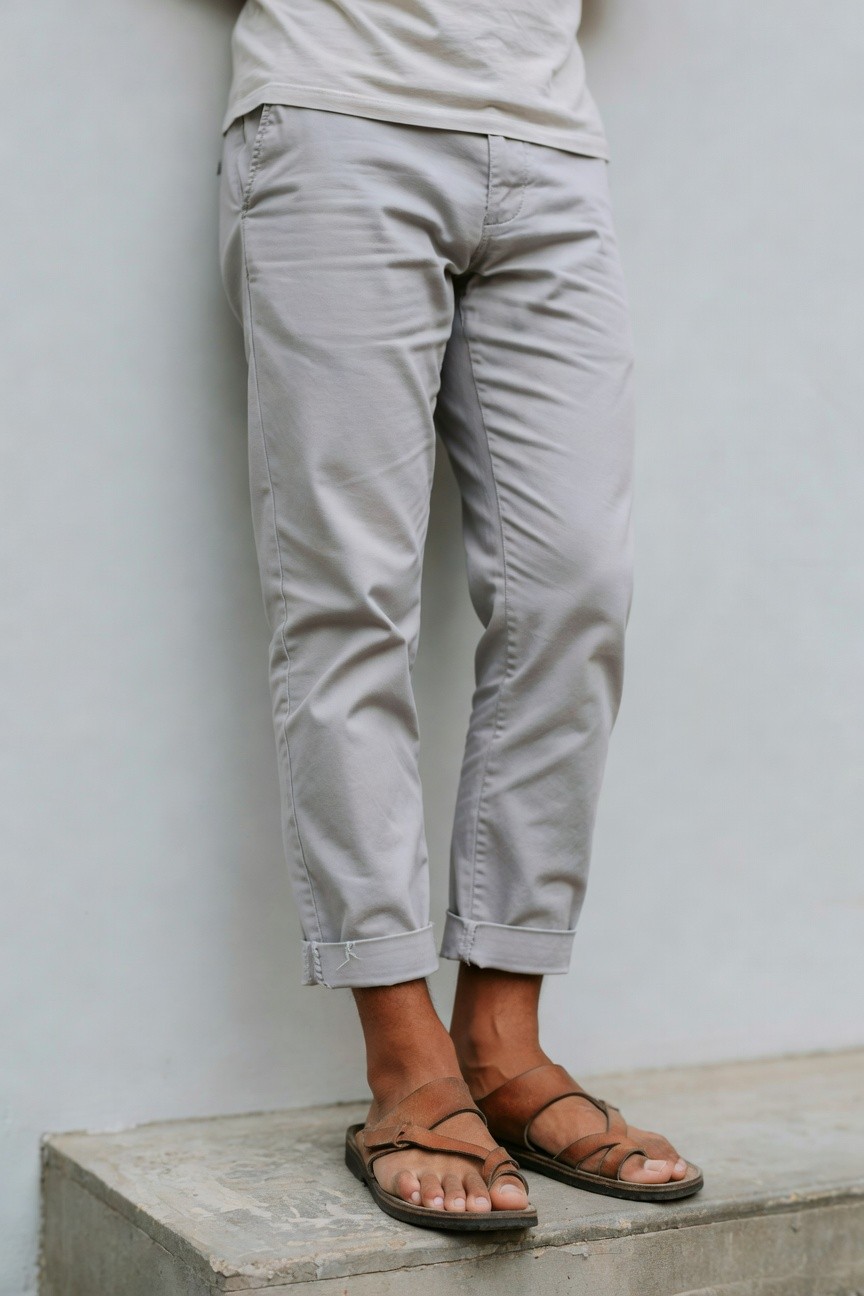 Man in light grey cropped chinos, beige t-shirt, and brown strappy leather sandals standing casually against white wall