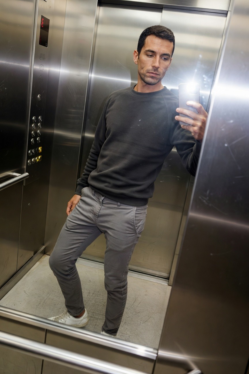 Man taking elevator mirror selfie in grey crewneck sweatshirt, slim grey pants, and white sneakers for a minimalist layered look
