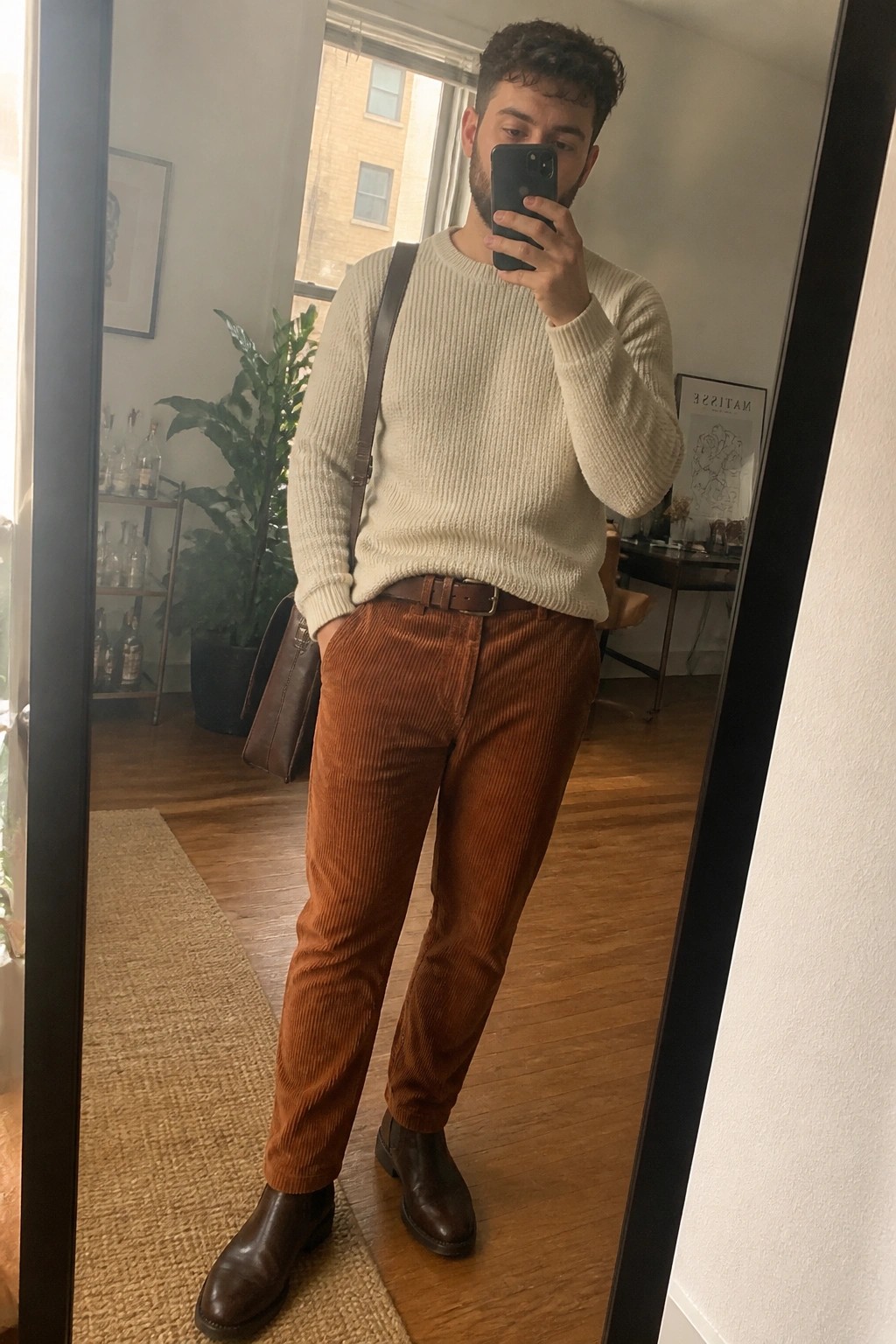 Man in cream sweater and brown corduroy pants taking mirror selfie in apartment