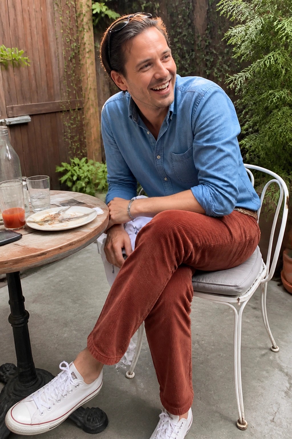 Smiling man in blue denim shirt and rust corduroy pants at outdoor table.