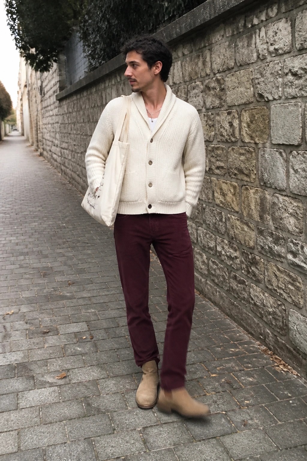 Man in cream cardigan and maroon pants with tote bag on cobblestone path.