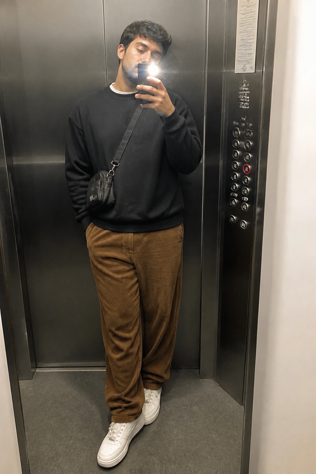 Man wearing brown corduroy pants, black sweatshirt, white sneakers and small black crossbody bag standing in an elevator while taking a mirror selfie.