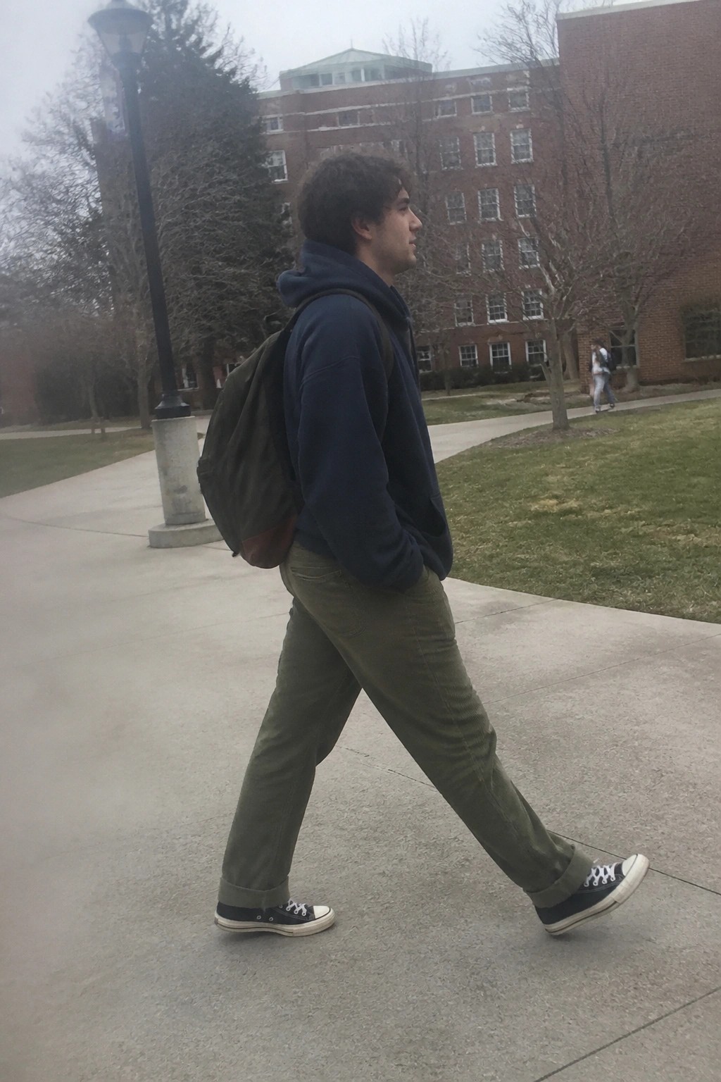 Young man in navy hoodie and green corduroy pants walks with backpack on campus path