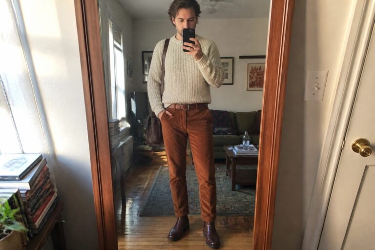 Man in cream sweater and brown corduroy pants taking mirror selfie in apartment