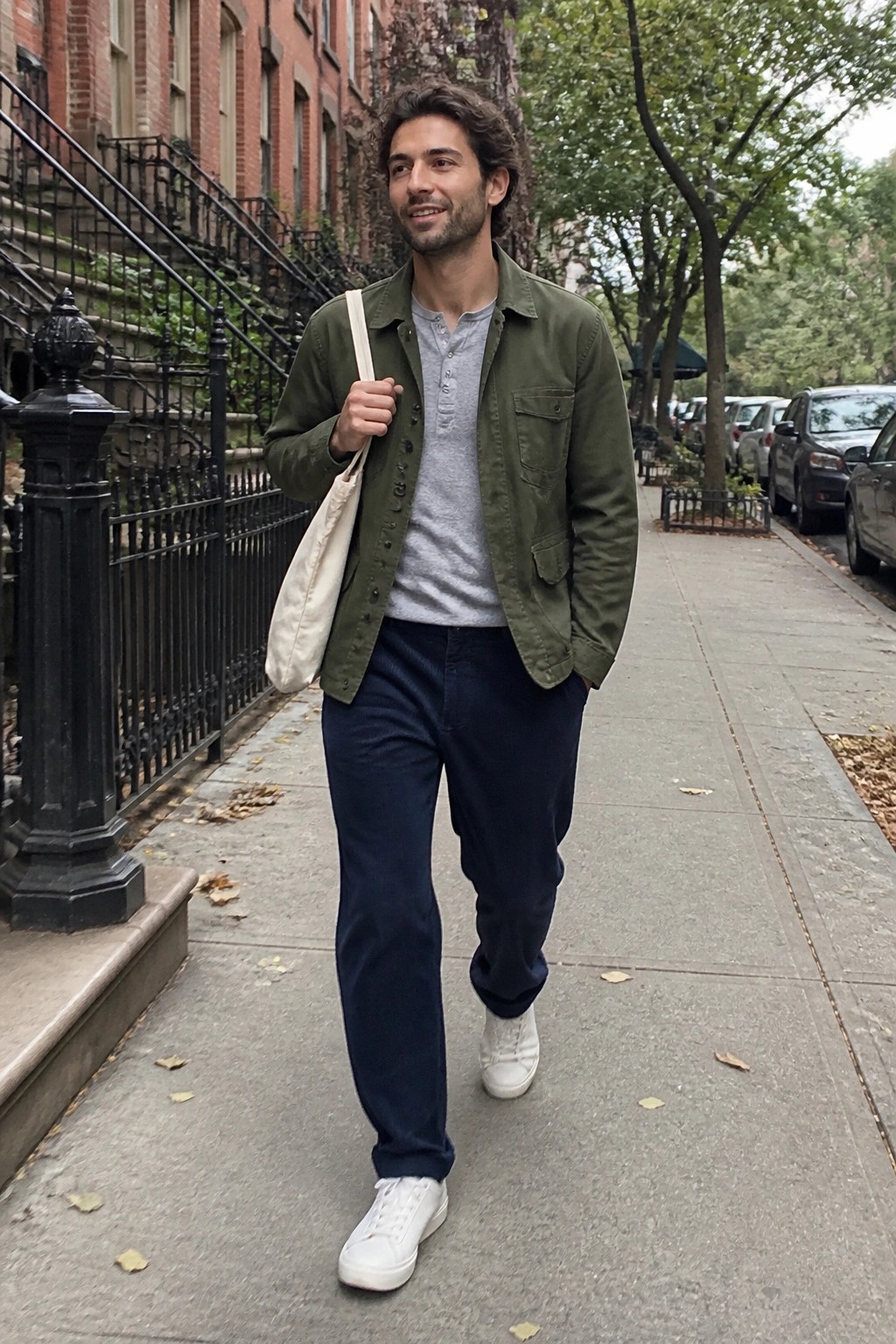 Man in green jacket carries tote bag while walking on city sidewalk