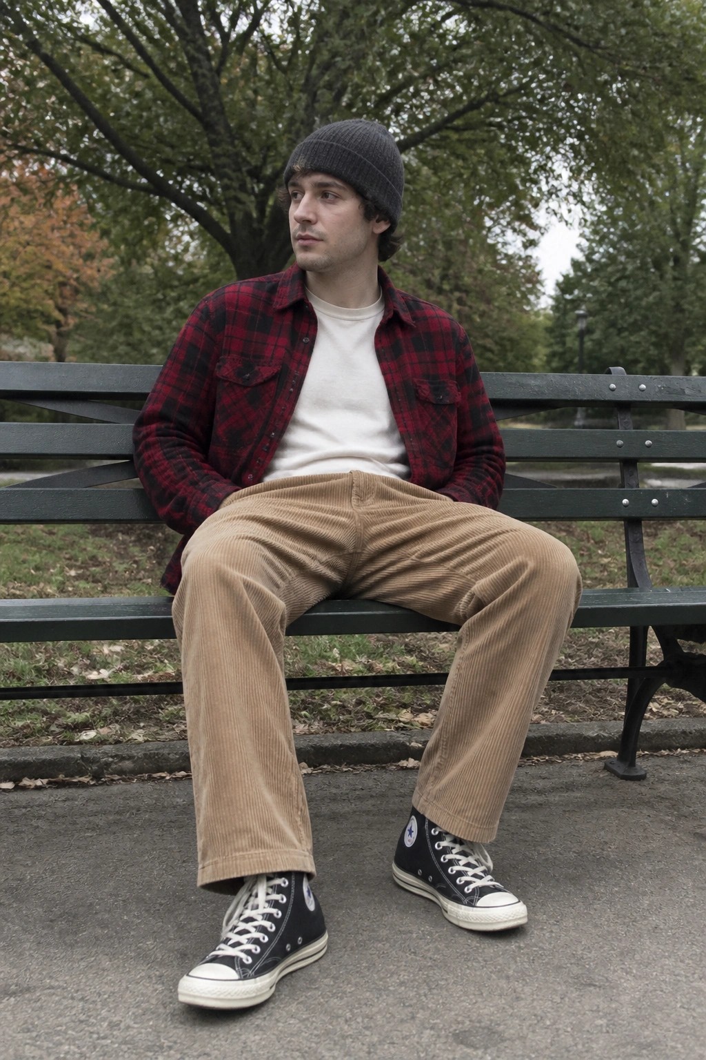 Man seated on a park bench wearing brown corduroy pants, a red and black plaid flannel shirt over a white t-shirt, black converse sneakers, and a gray beanie.