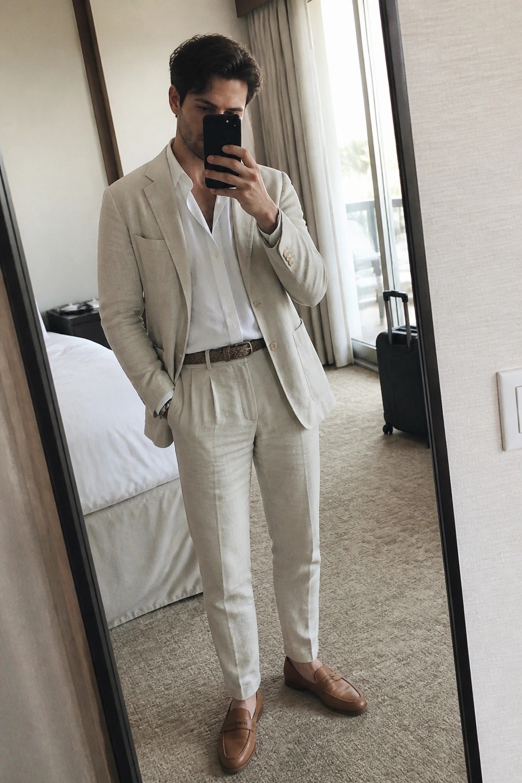 Man in beige linen suit and white shirt taking mirror selfie in hotel room