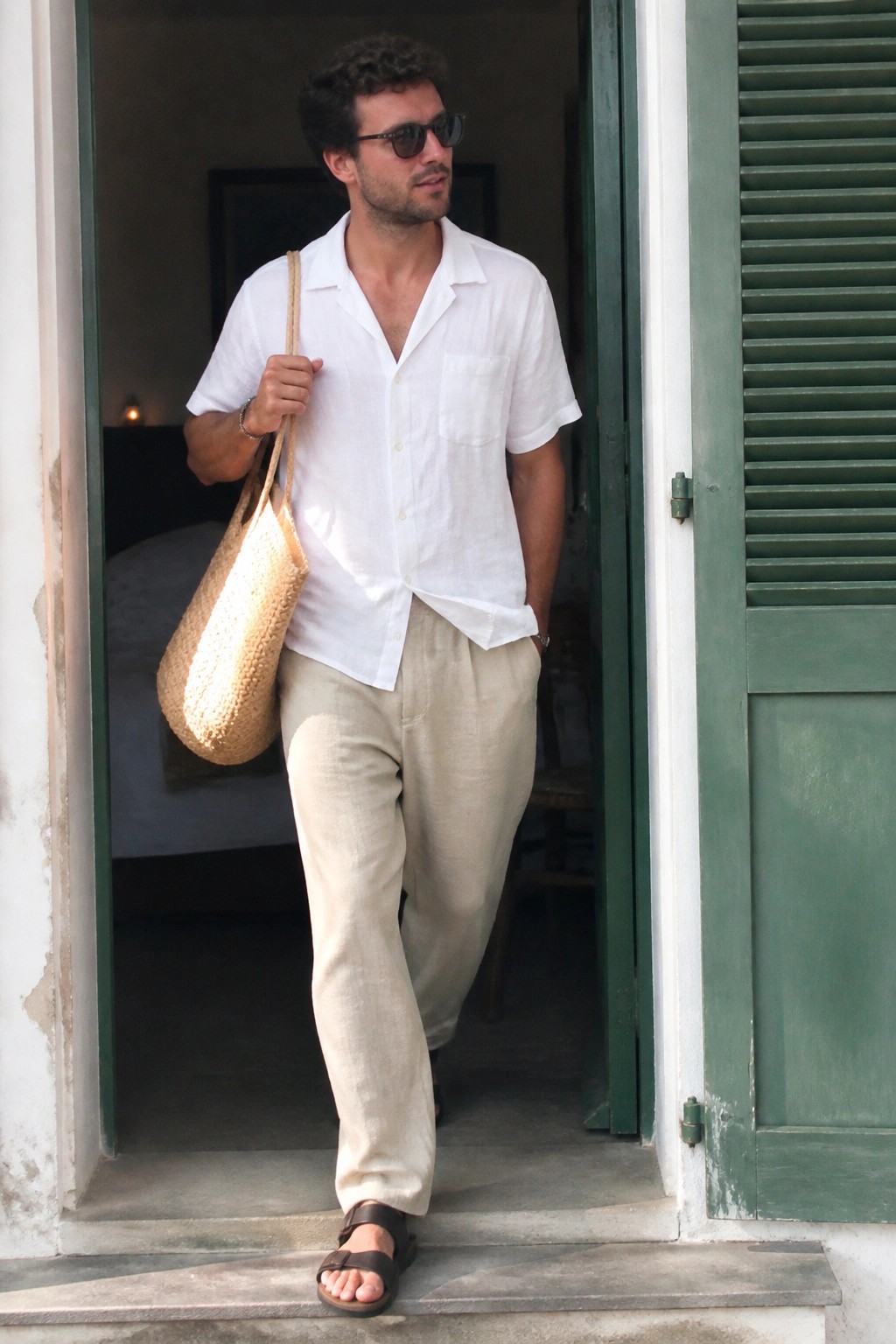 Man in white shirt and beige pants carries straw bag exiting green doorway