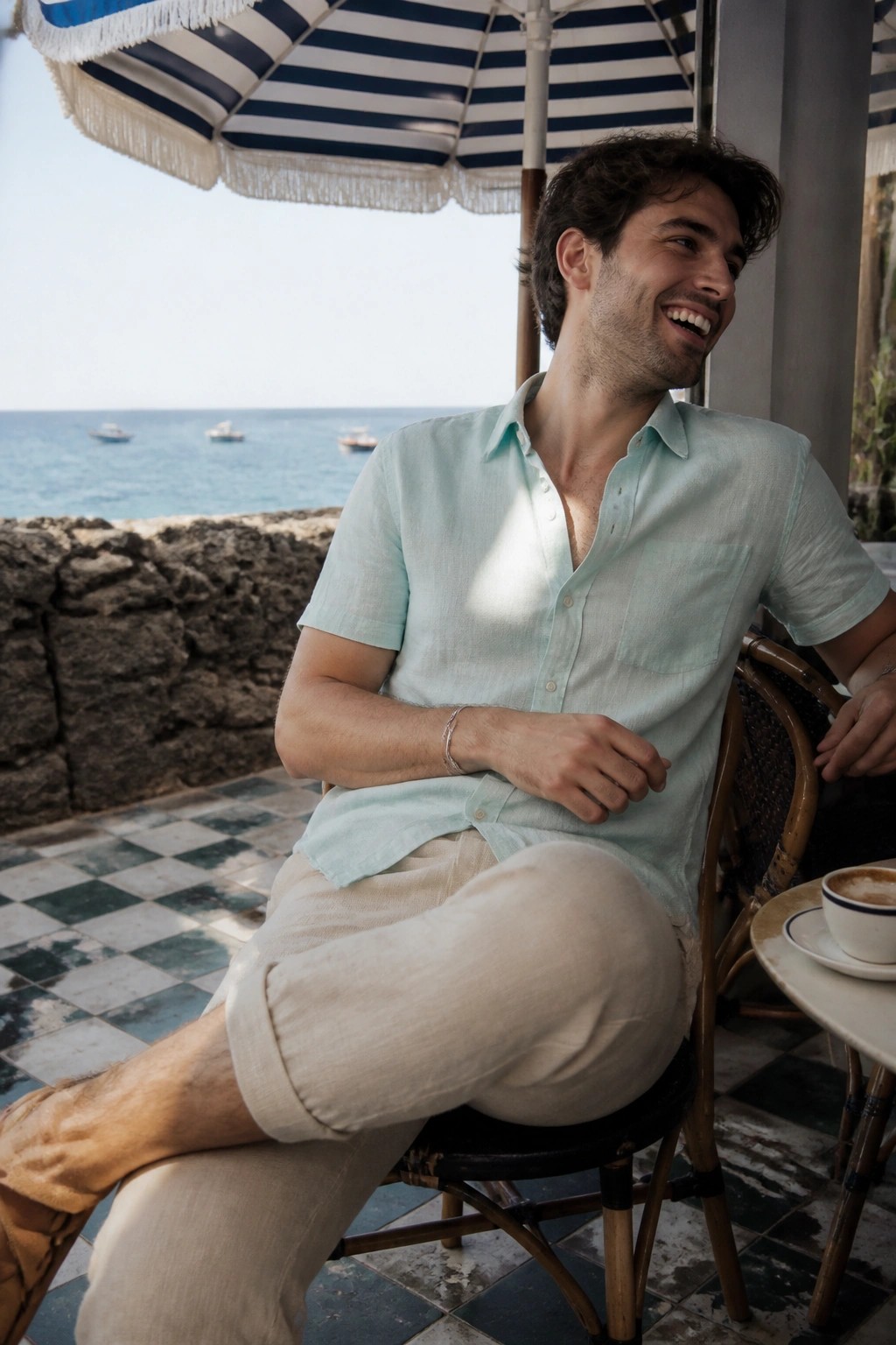 Smiling man in mint shirt and beige pants at seaside cafe under striped umbrella.