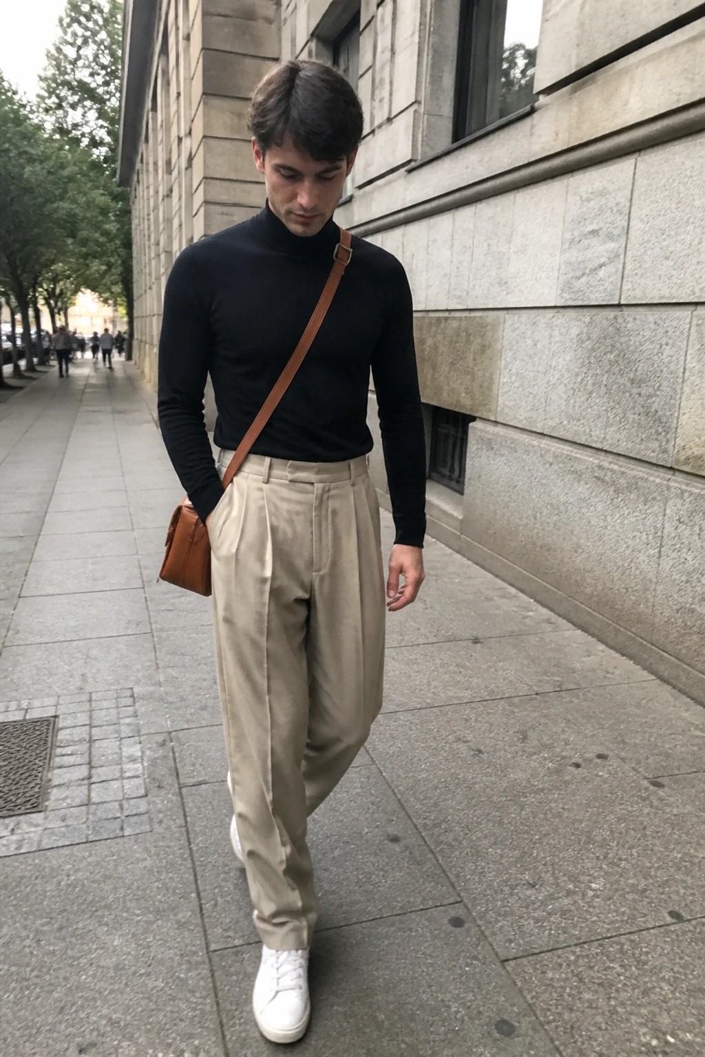 A man wearing beige pleated formal pants, black turtleneck sweater, white sneakers, and brown crossbody bag walking on a city sidewalk.