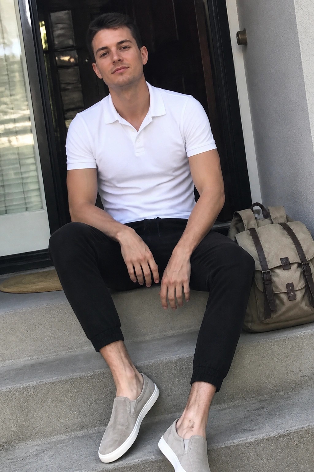 Man in white polo shirt and black pants sitting on steps beside backpack