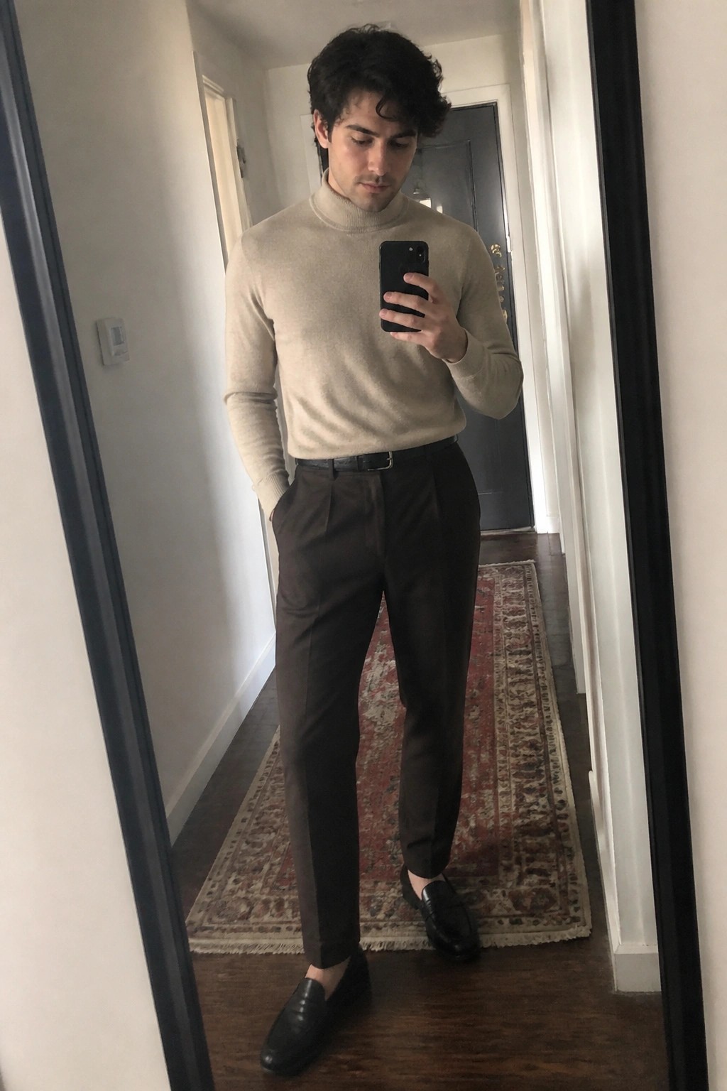 Man in beige turtleneck and brown trousers taking mirror selfie in hallway