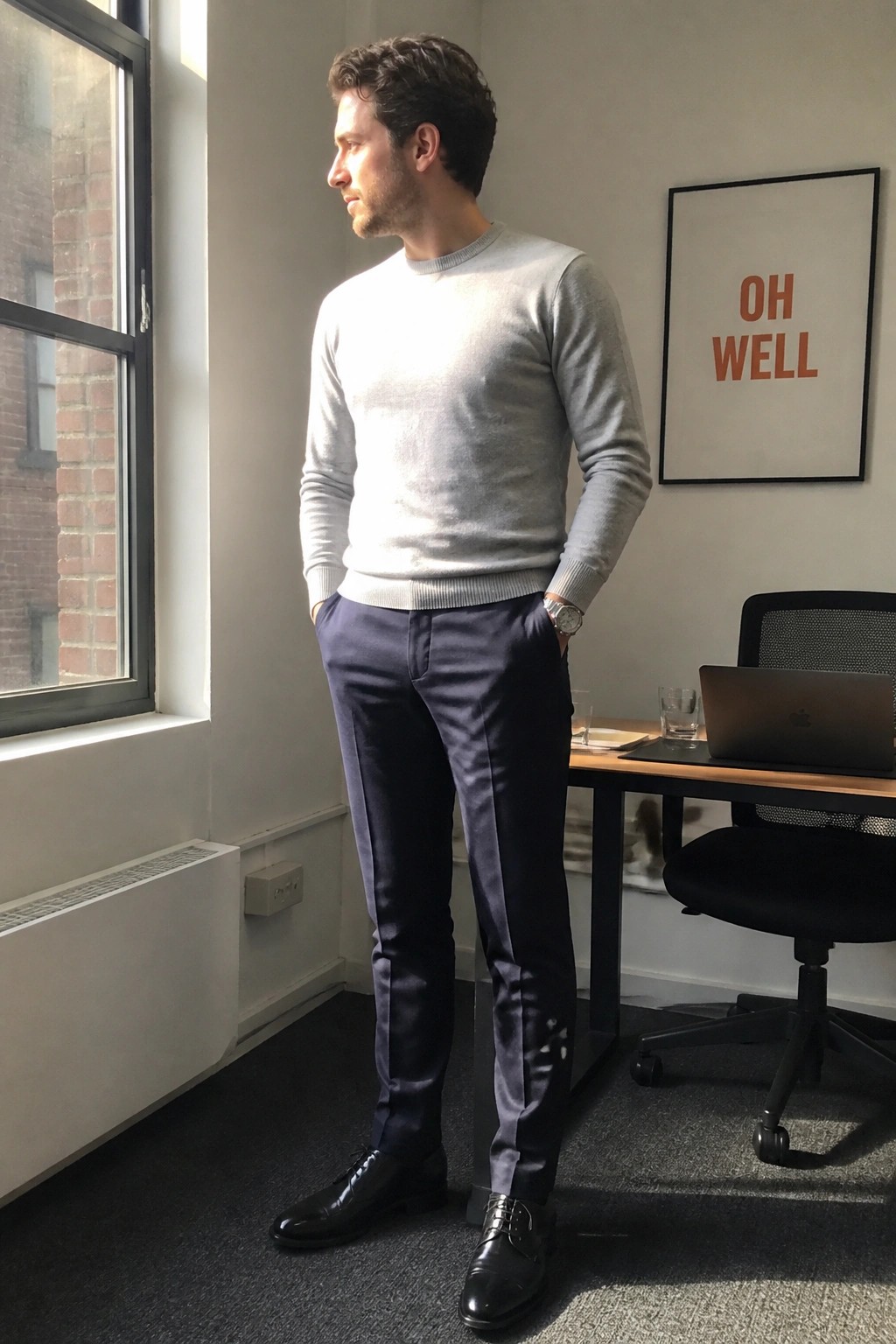 Man in gray sweater and navy pants standing in sunlit modern office by window