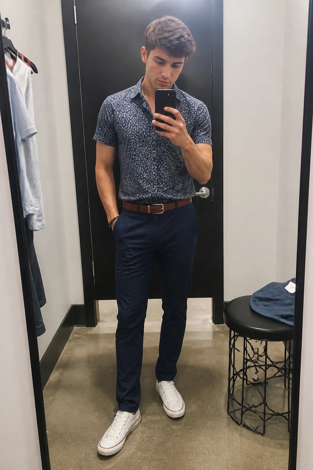 Young man in patterned shirt and navy pants taking mirror selfie in fitting room.