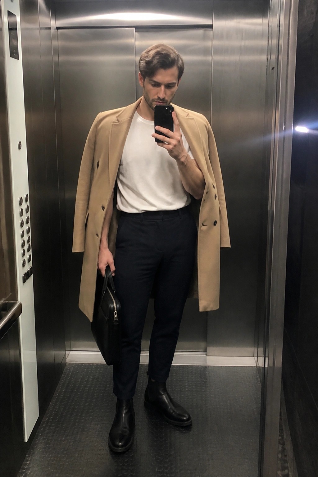 Man in tan overcoat taking mirror selfie in elevator while holding black bag